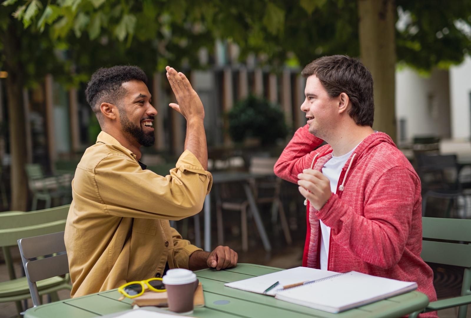 Two friends laughing and high-fiving at an outdoor café table — one of them has Down syndrome